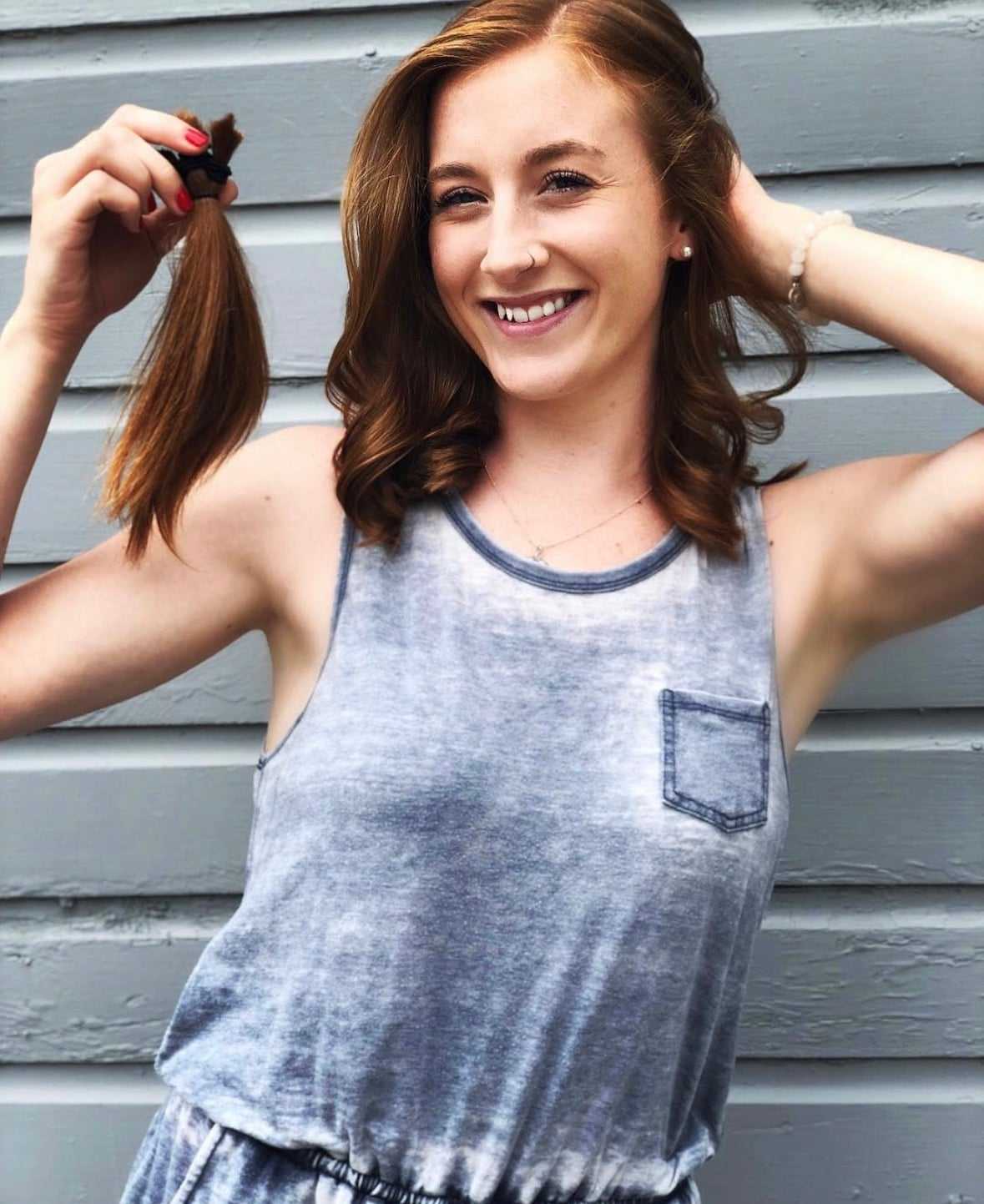 Woman smiling, holding cut hair in hand, standing against a gray wall.